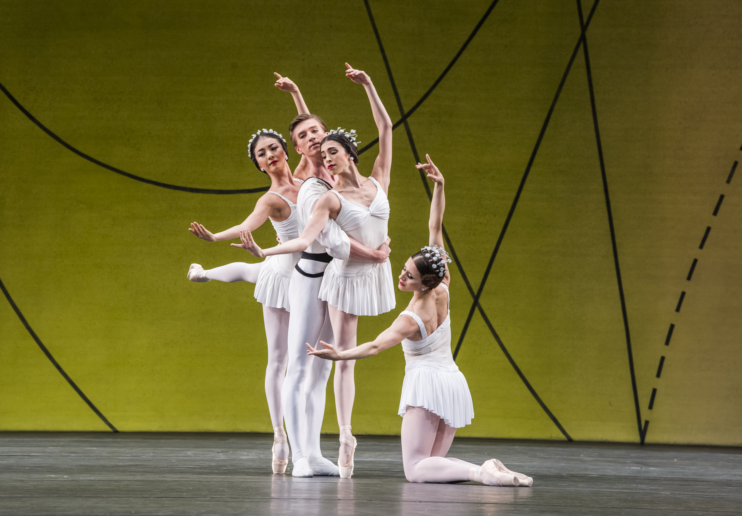 Several ballet dancers perform on stage, all are wearing white costumes. They perform in front of a yellow and green gradient backdrop with flowing geometric patterns. (4 of 6)
