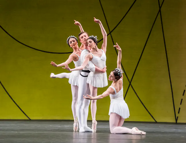 Several ballet dancers perform on stage, all are wearing white costumes. They perform in front of a yellow and green gradient backdrop with flowing geometric patterns. (4 of 6)