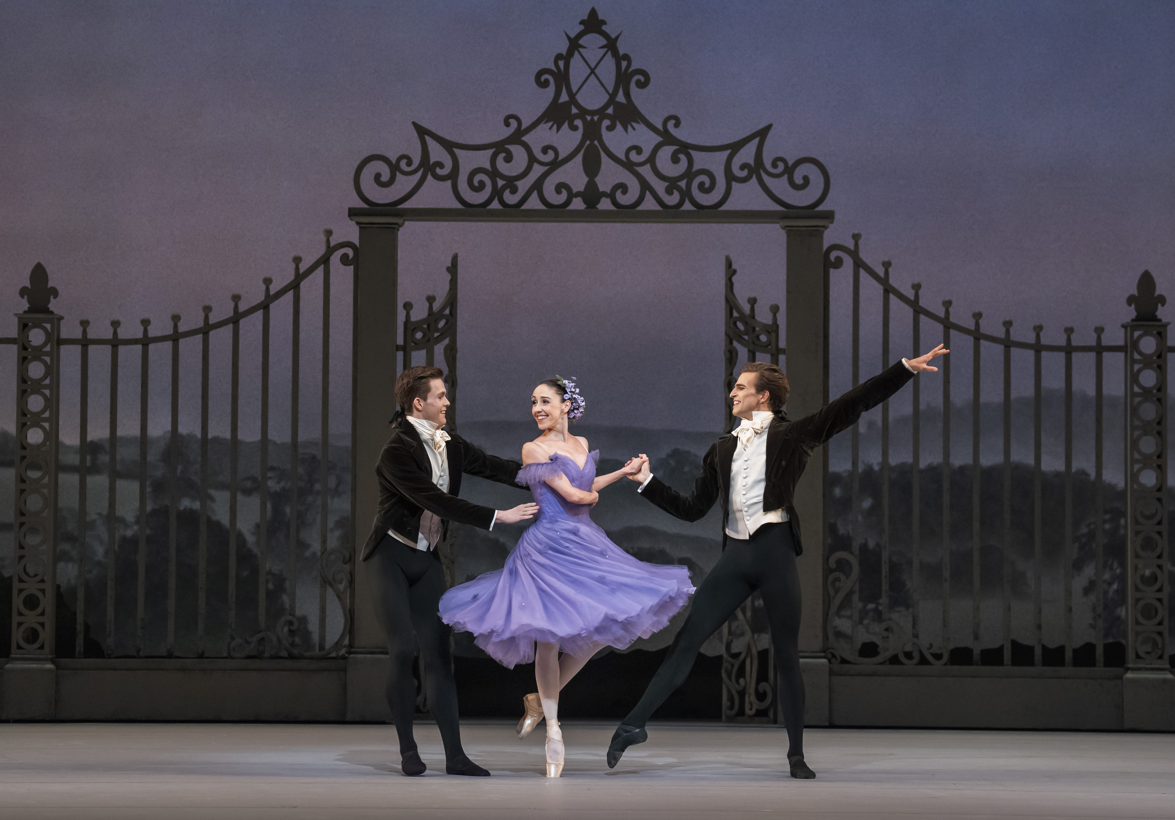 Two ballet dancers wearing black tuxedo jackets and white dress shirts perform with a ballet dancer wearing a purple dress with flowers in her hair. They perform in front of an ornate gate with a painterly rural background. (2 of 6)