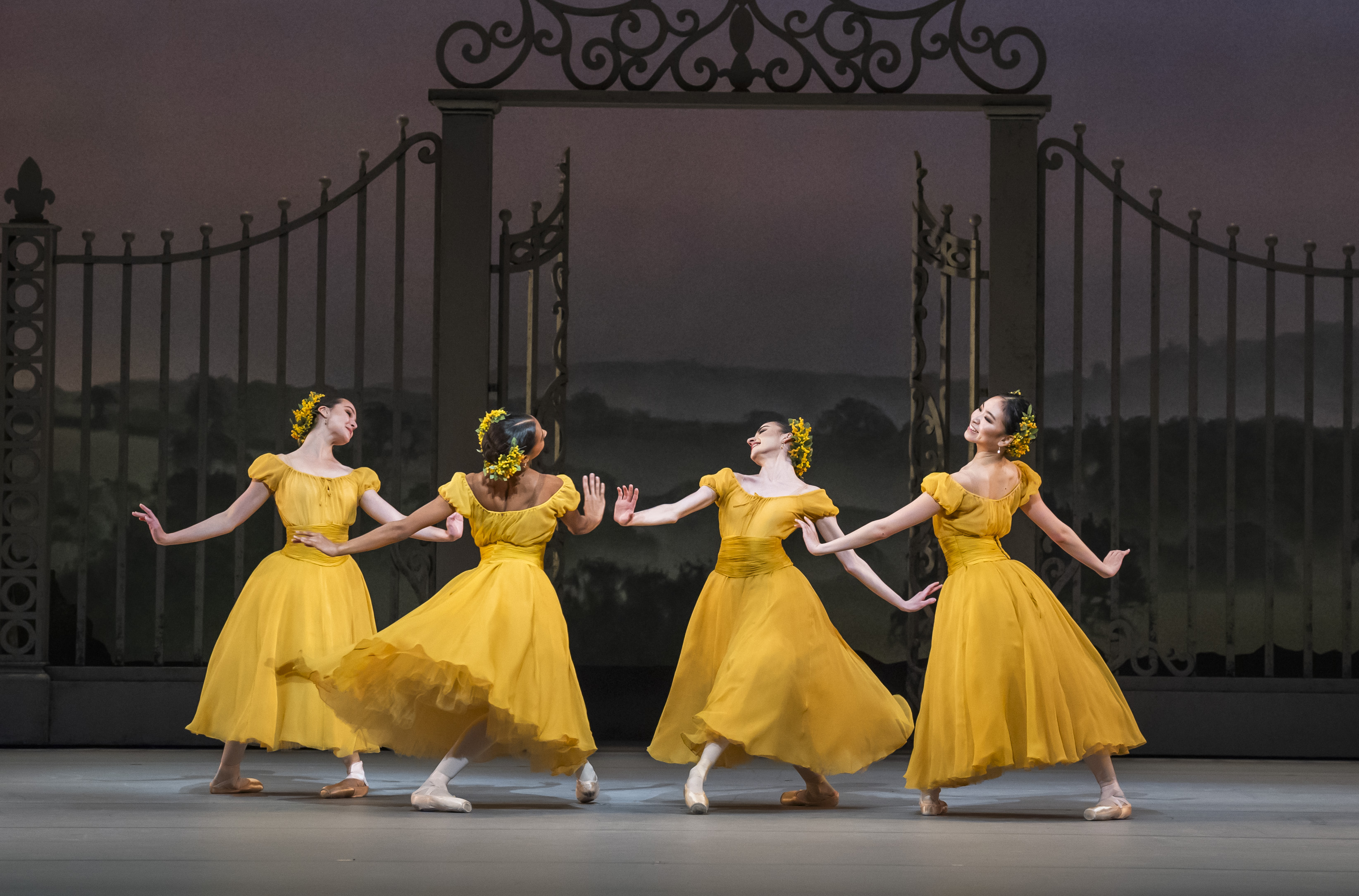 Four ballet dancers wearing yellow dresses with flowers in their hair perform together in the centre of a stage. They perform in front of an ornate gate with a painterly rural background.