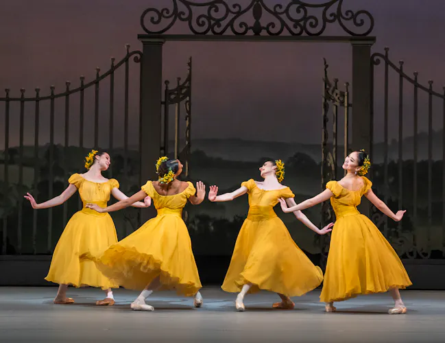 Four ballet dancers wearing yellow dresses with flowers in their hair perform together in the centre of a stage. They perform in front of an ornate gate with a painterly rural background. (1 of 6)