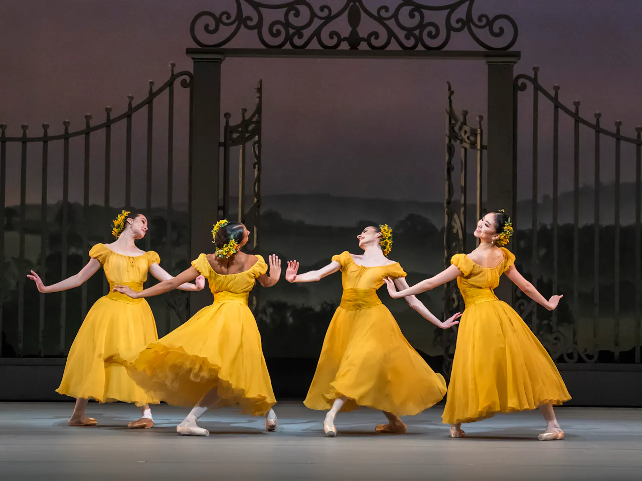 Four ballet dancers wearing yellow dresses with flowers in their hair perform together in the centre of a stage. They perform in front of an ornate gate with a painterly rural background.