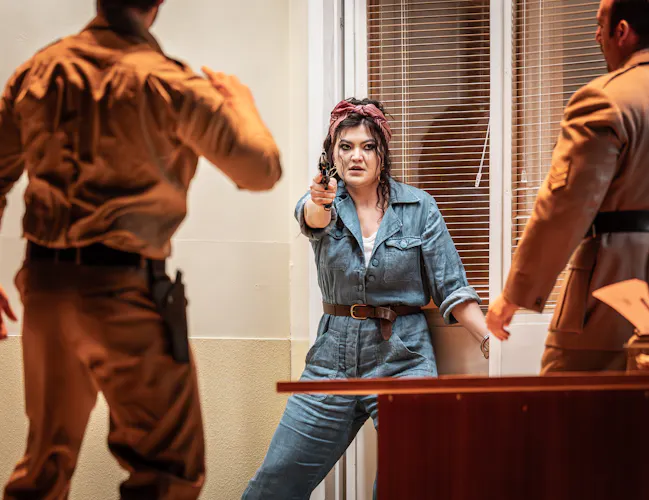 A performer wearing a blue denim jumpsuit and bandana hair scarf holds a gun towards two other performers with their backs to the audience. They are performing in The Royal Opera's production of Carmen. (1 of 7)