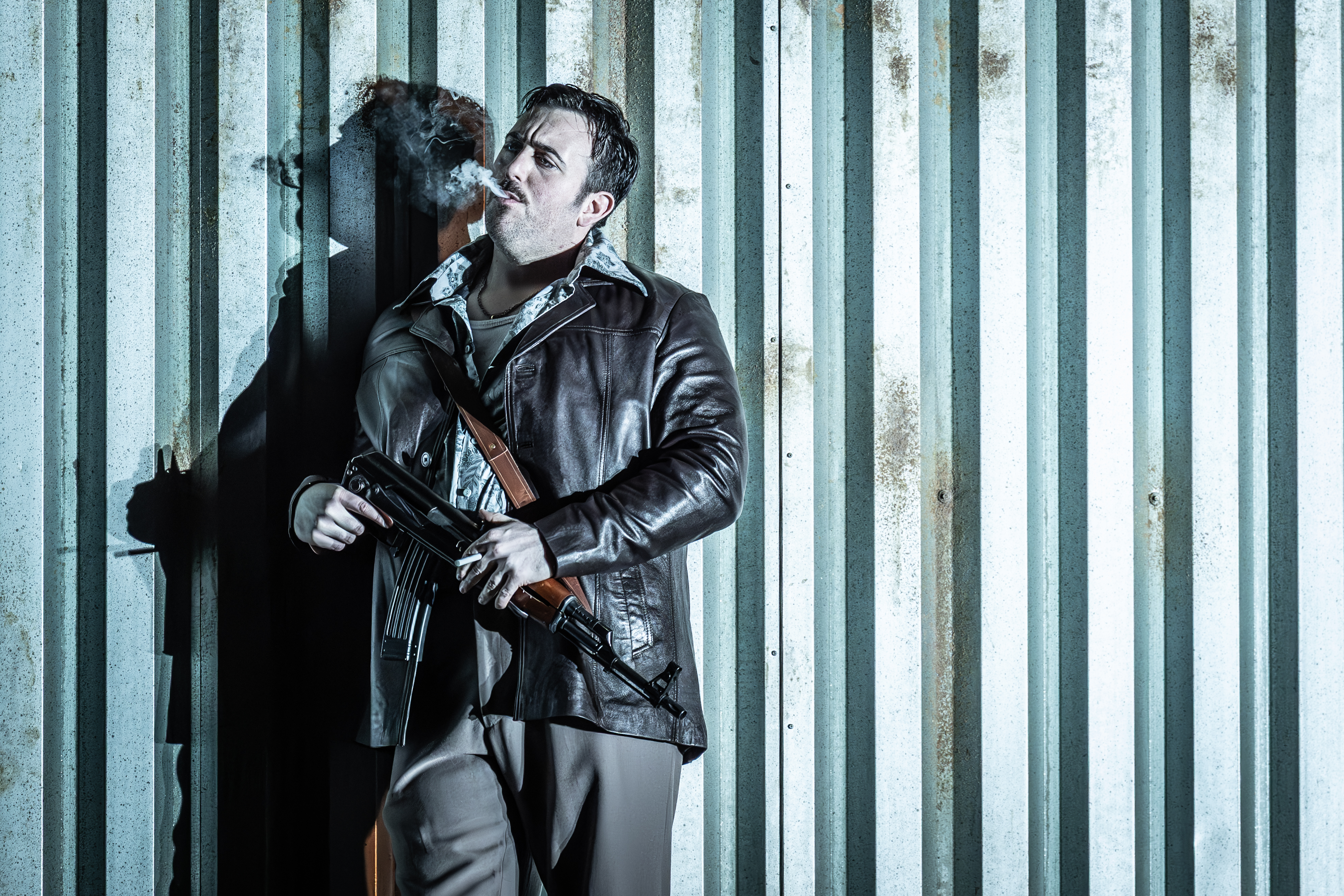 A performer wearing a leather jacket stands agains a corrugated iron wall smoking a cigarette. They are performing in The Royal Opera's production of Carmen. (2 of 7)