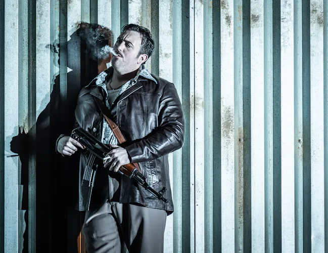 A performer wearing a leather jacket stands agains a corrugated iron wall smoking a cigarette. They are performing in The Royal Opera's production of Carmen. (2 of 7)