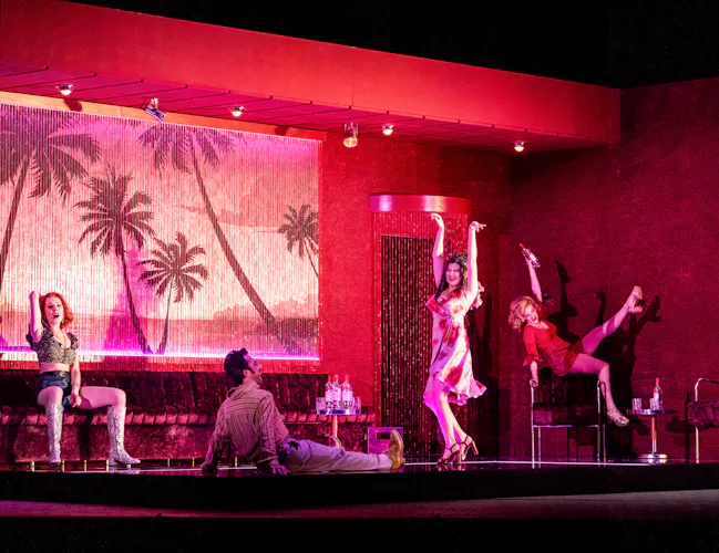 A bar scene on a stage with pink and red neon lighting and several performers dancing. They are performing in The Royal Opera's production of Carmen. (7 of 7)