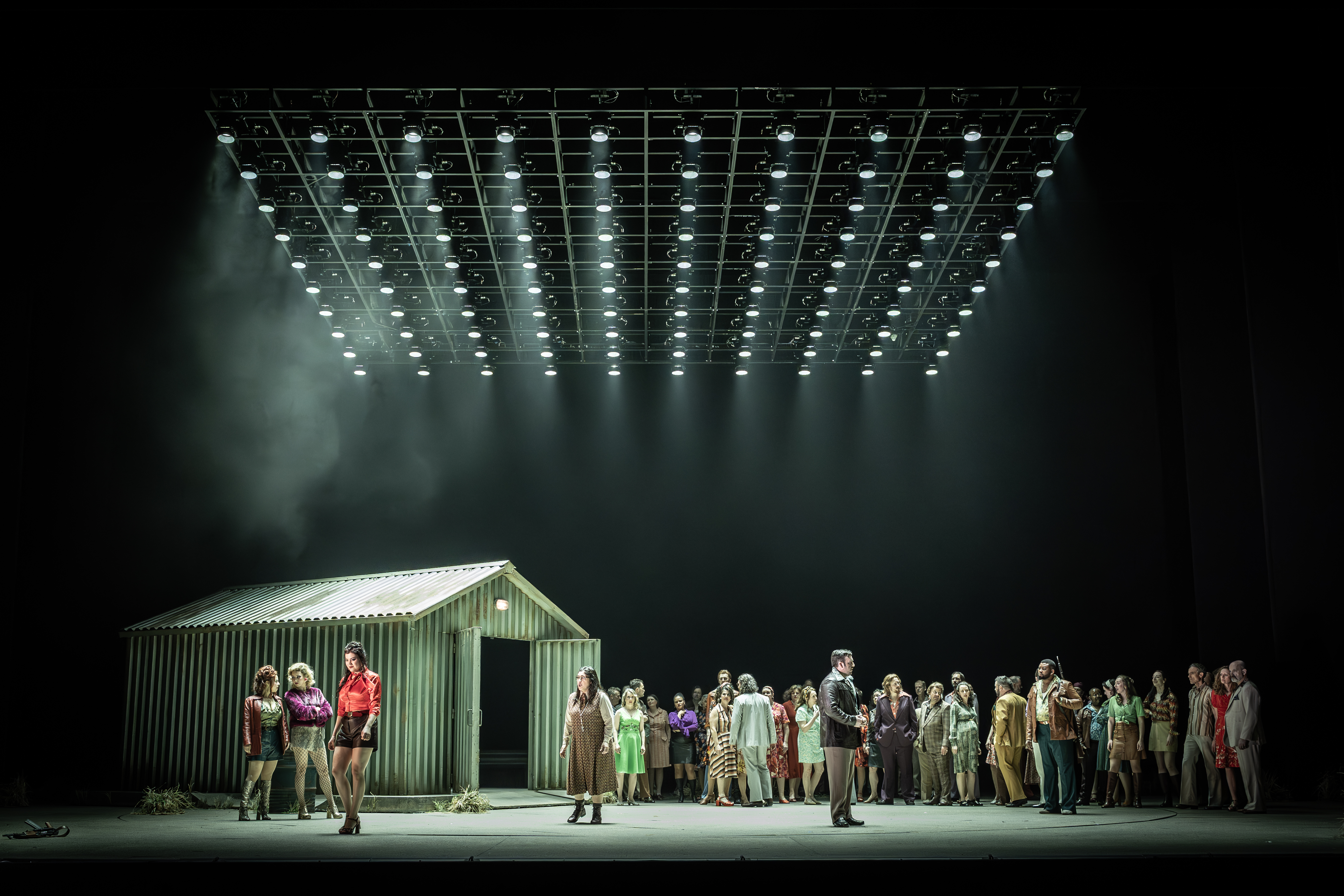 A corrugated iron shed sits on stage. A group of brightly dressed performers stands alongside it. The stage is dimly lit from above from a rig of lighting. They are performing in The Royal Opera's production of Carmen. (4 of 7)