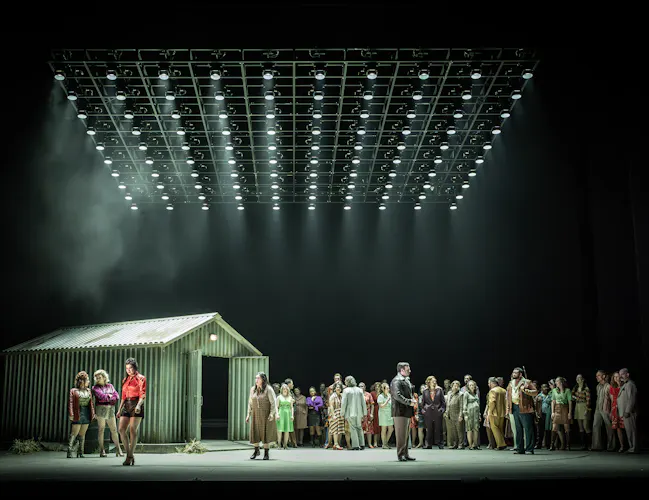 A corrugated iron shed sits on stage. A group of brightly dressed performers stands alongside it. The stage is dimly lit from above from a rig of lighting. They are performing in The Royal Opera's production of Carmen. (4 of 7)
