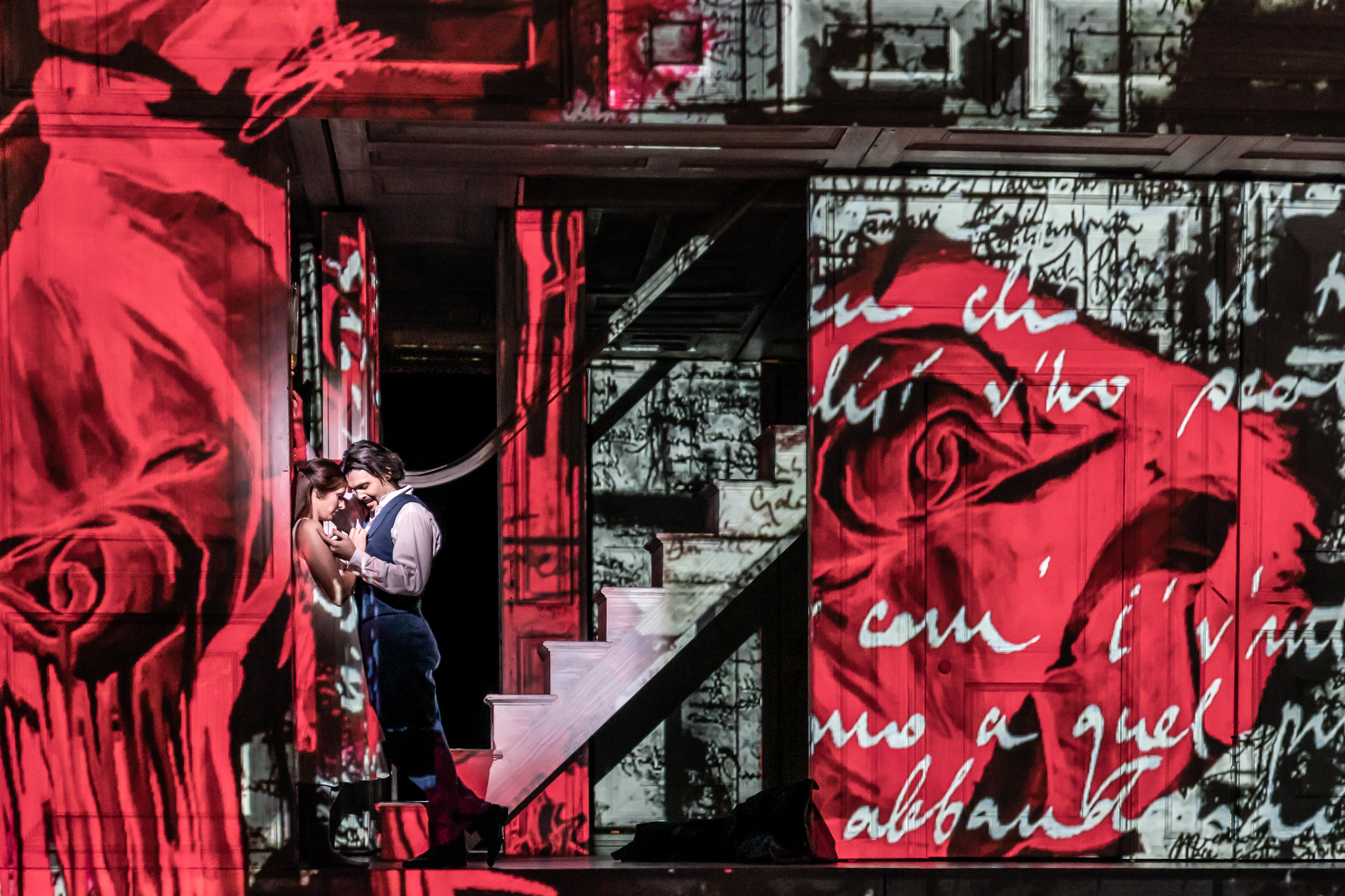 Two performers embrace passionately on a stage in front of a backdrop of a house that has images of red roses and handwriting projected onto the entire space. 