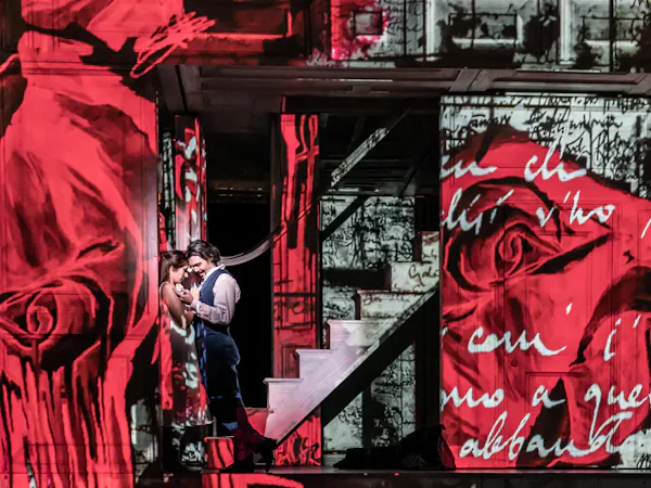 Two performers embrace passionately on a stage in front of a backdrop of a house that has images of red roses and handwriting projected onto the entire space.