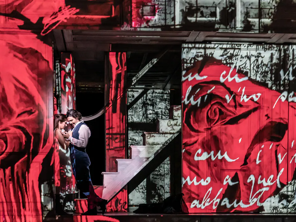 Two performers embrace passionately on a stage in front of a backdrop of a house that has images of red roses and handwriting projected onto the entire space.