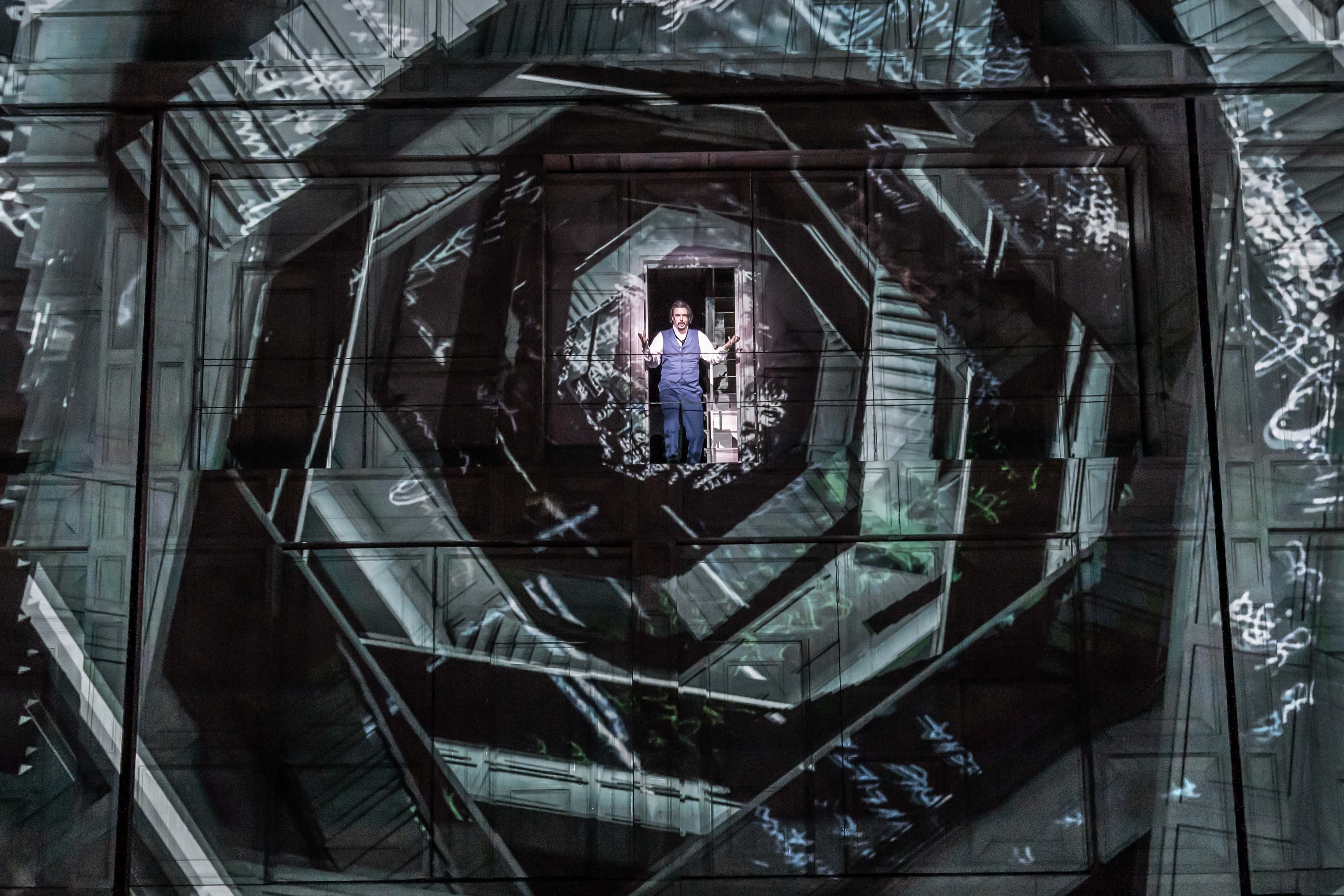 An opera singer wearing blue trousers and waistcoat and a white shirt stands in the centre of several layered geometric shapes that appear to have staircases set within. It is an artistic image from The Royal Opera's production of Don Giovanni. (1 of 4)