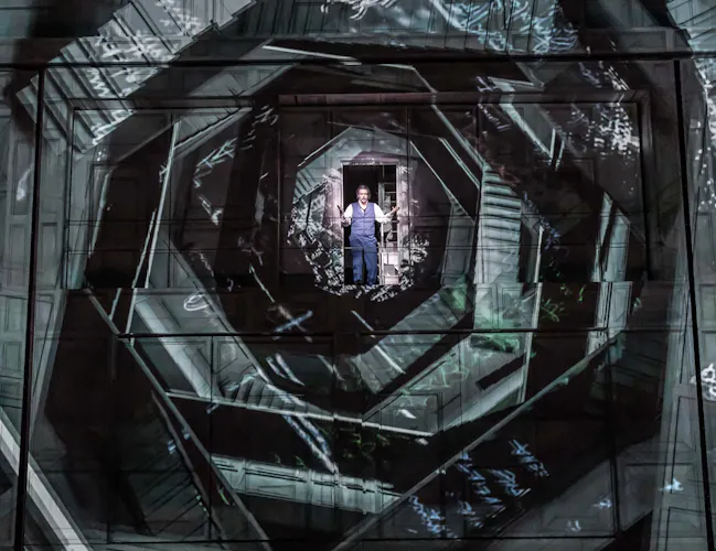 An opera singer wearing blue trousers and waistcoat and a white shirt stands in the centre of several layered geometric shapes that appear to have staircases set within. It is an artistic image from The Royal Opera's production of Don Giovanni. (1 of 4)