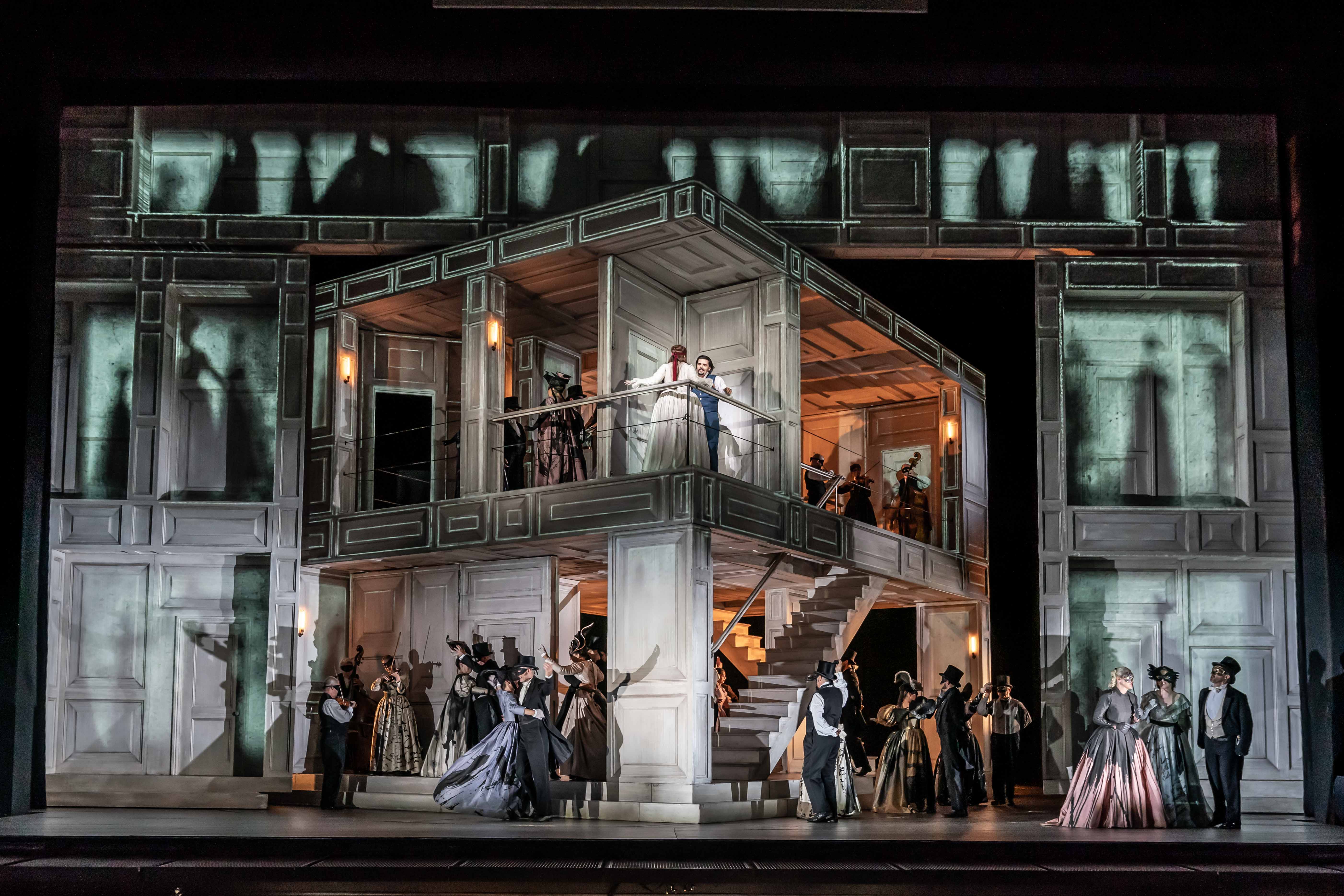 An open view of the interior of a house with two floors which stands in front of a backdrop of another building with brightly lit windows with the shadows of people. Several performers are inside the house on both floors. It is from The Royal Opera's production of Don Giovanni.m (2 of 4)