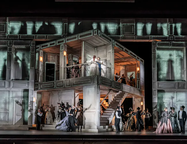 An open view of the interior of a house with two floors which stands in front of a backdrop of another building with brightly lit windows with the shadows of people. Several performers are inside the house on both floors. It is from The Royal Opera's production of Don Giovanni.m (2 of 4)