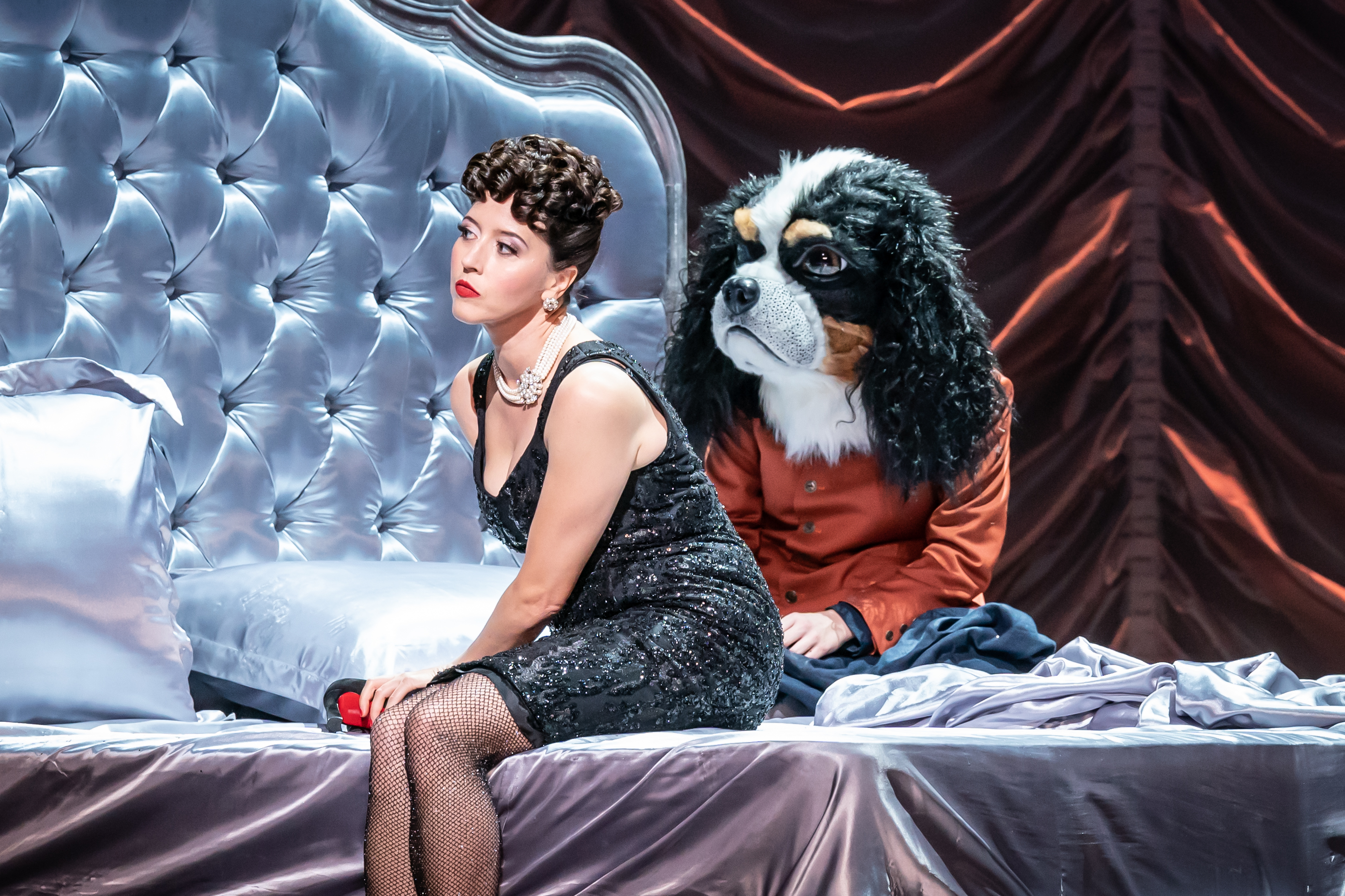 A performer wearing a glamorous black dress, fishnet tights and a chunky pearl necklace sits on a large silk dressed bed. Next to them sits a performer wearing an orange jacket and a mask of a King Charles spaniel. They are on stage of The Royal Opera House in a Royal Opera production of Alcina. (3 of 6)