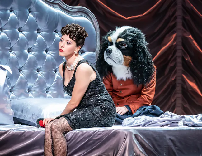 A performer wearing a glamorous black dress, fishnet tights and a chunky pearl necklace sits on a large silk dressed bed. Next to them sits a performer wearing an orange jacket and a mask of a King Charles spaniel. They are on stage of The Royal Opera House in a Royal Opera production of Alcina. (3 of 6)