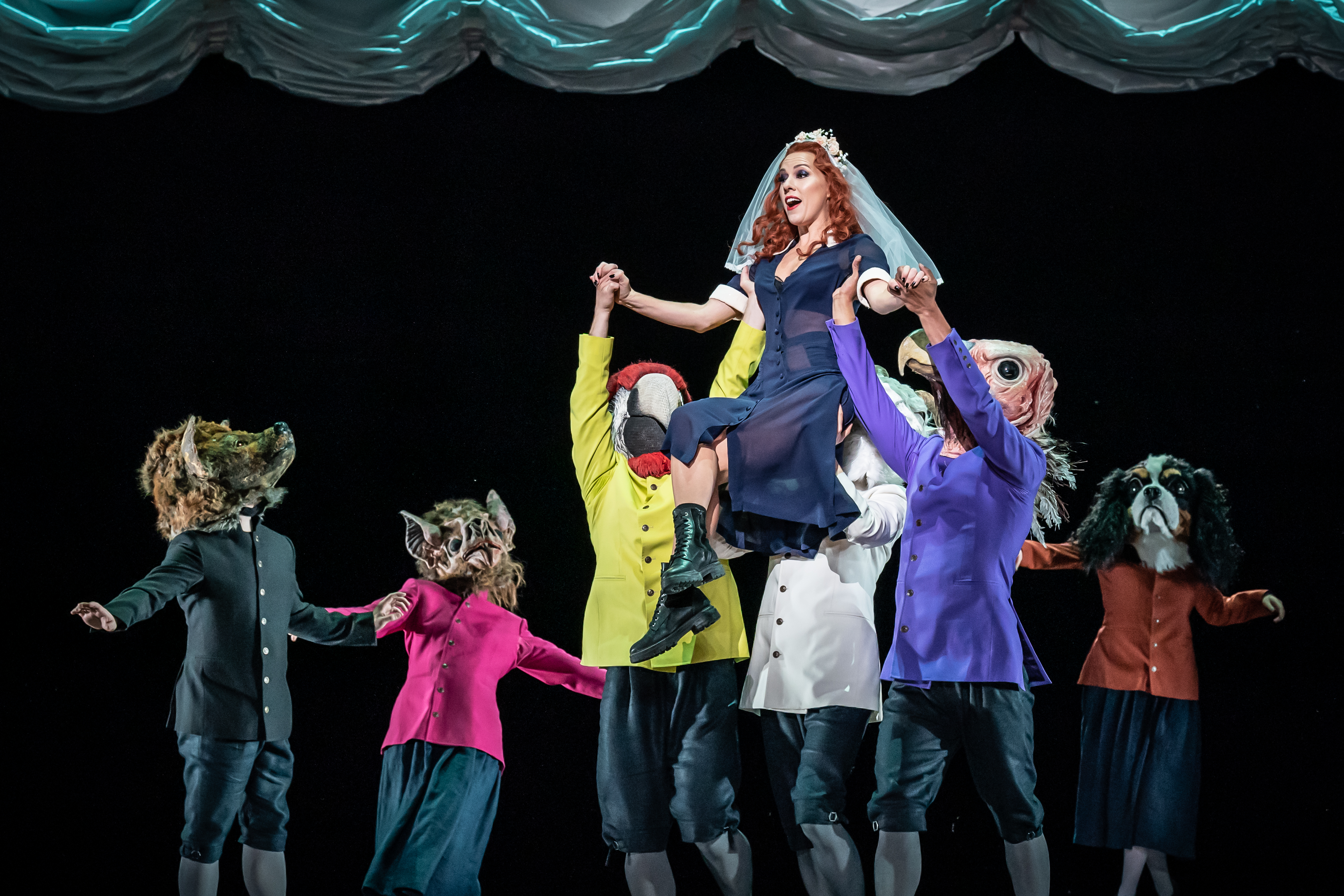 A performer wearing a black sheer dress, boots and a white veil is held up in the air by several performers wearing bright colour jackets and animal masks on stage. (1 of 6)