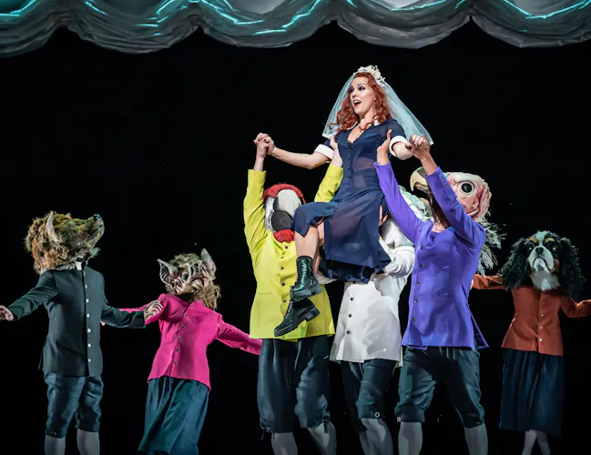 A performer wearing a black sheer dress, boots and a white veil is held up in the air by several performers wearing bright colour jackets and animal masks on stage. (1 of 6)