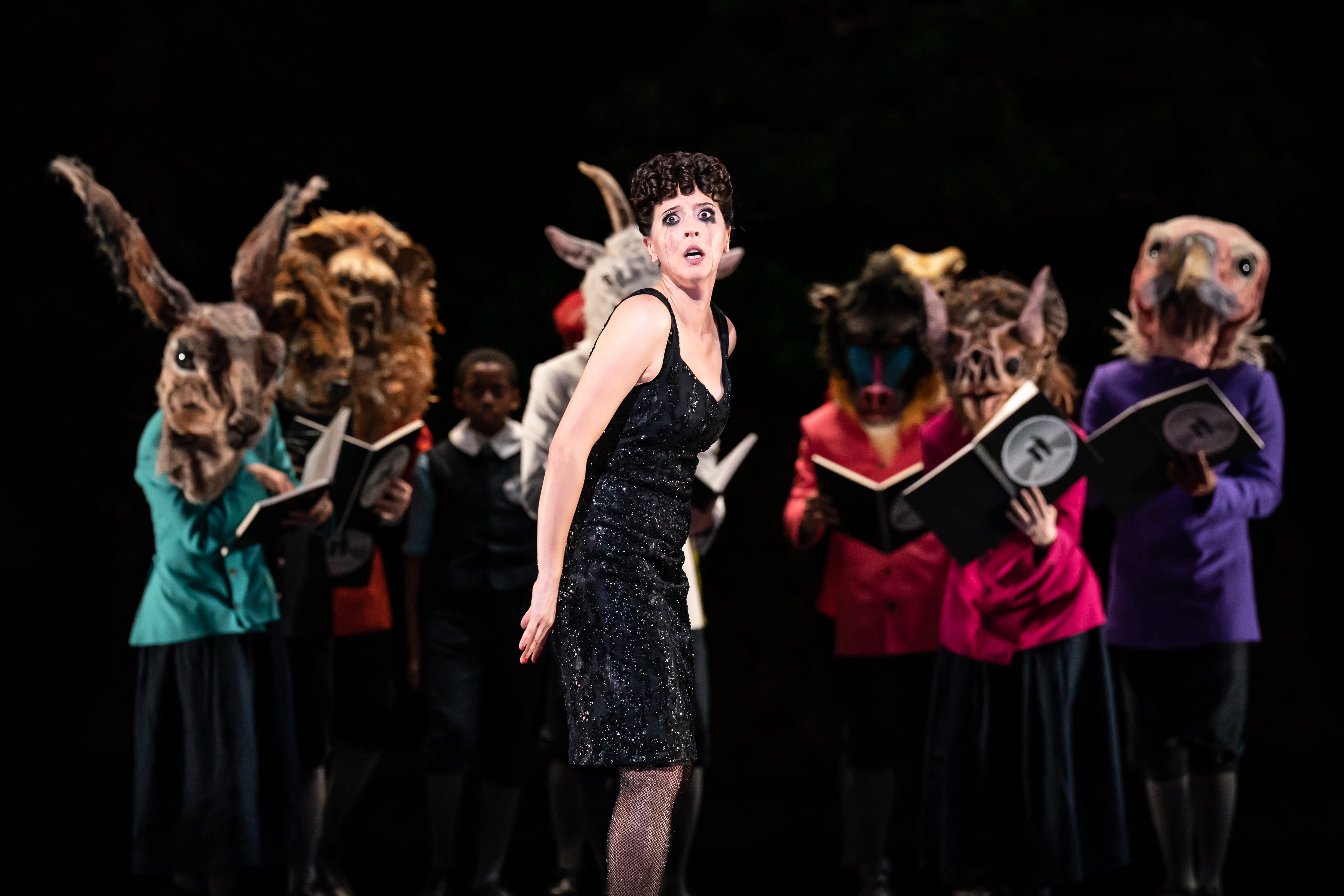 A performer wearing a glamorous black dress and fishnets stands on stage singing. Behind them are several performers wearing bright colour jackets and various animal masks, holding up books. (5 of 6)