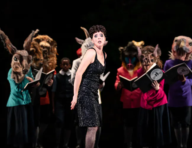 A performer wearing a glamorous black dress and fishnets stands on stage singing. Behind them are several performers wearing bright colour jackets and various animal masks, holding up books. (5 of 6)