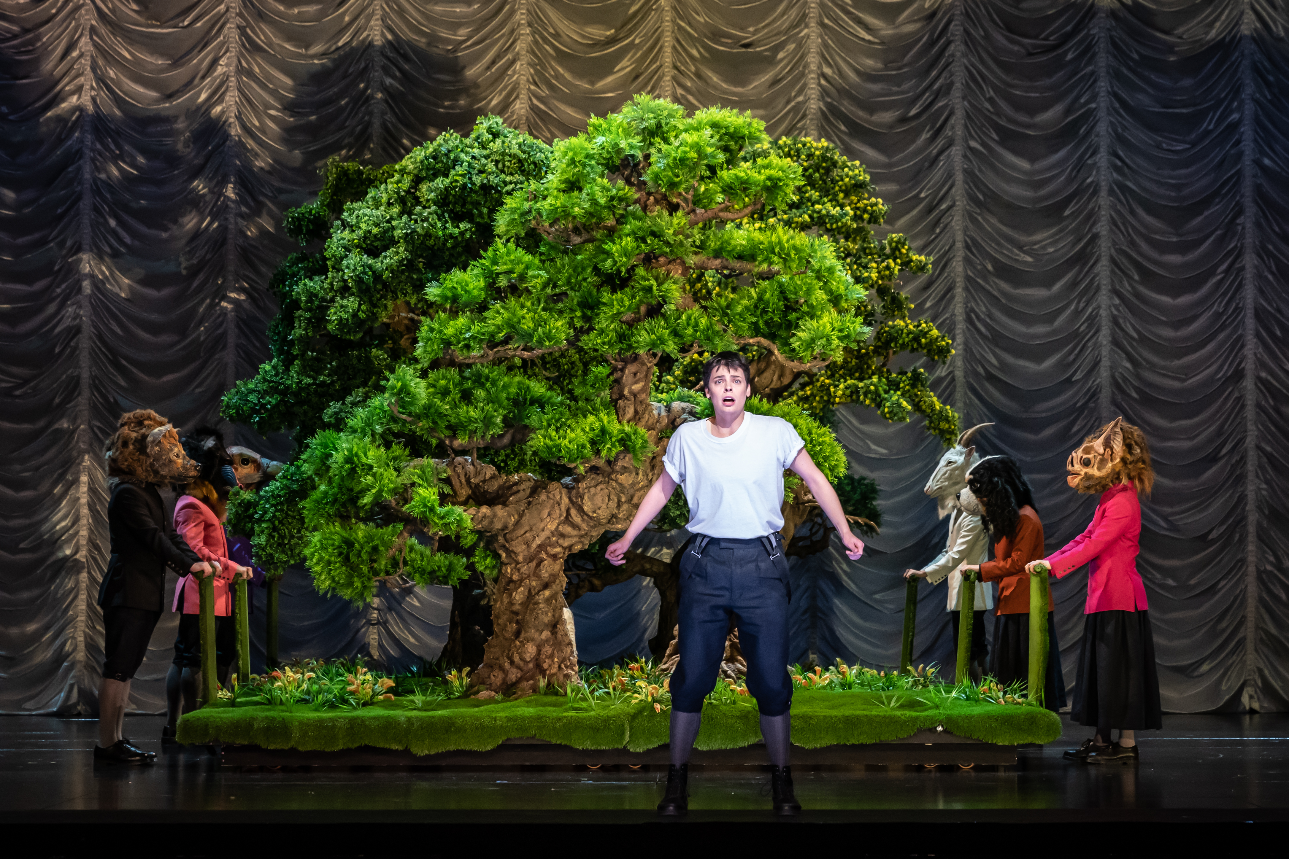 A performer stands in front of a small bonsai tree on stage. Either side of the bonsai stand performers wearing various animal masks. (6 of 6)