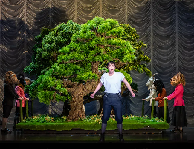 A performer stands in front of a small bonsai tree on stage. Either side of the bonsai stand performers wearing various animal masks. (6 of 6)