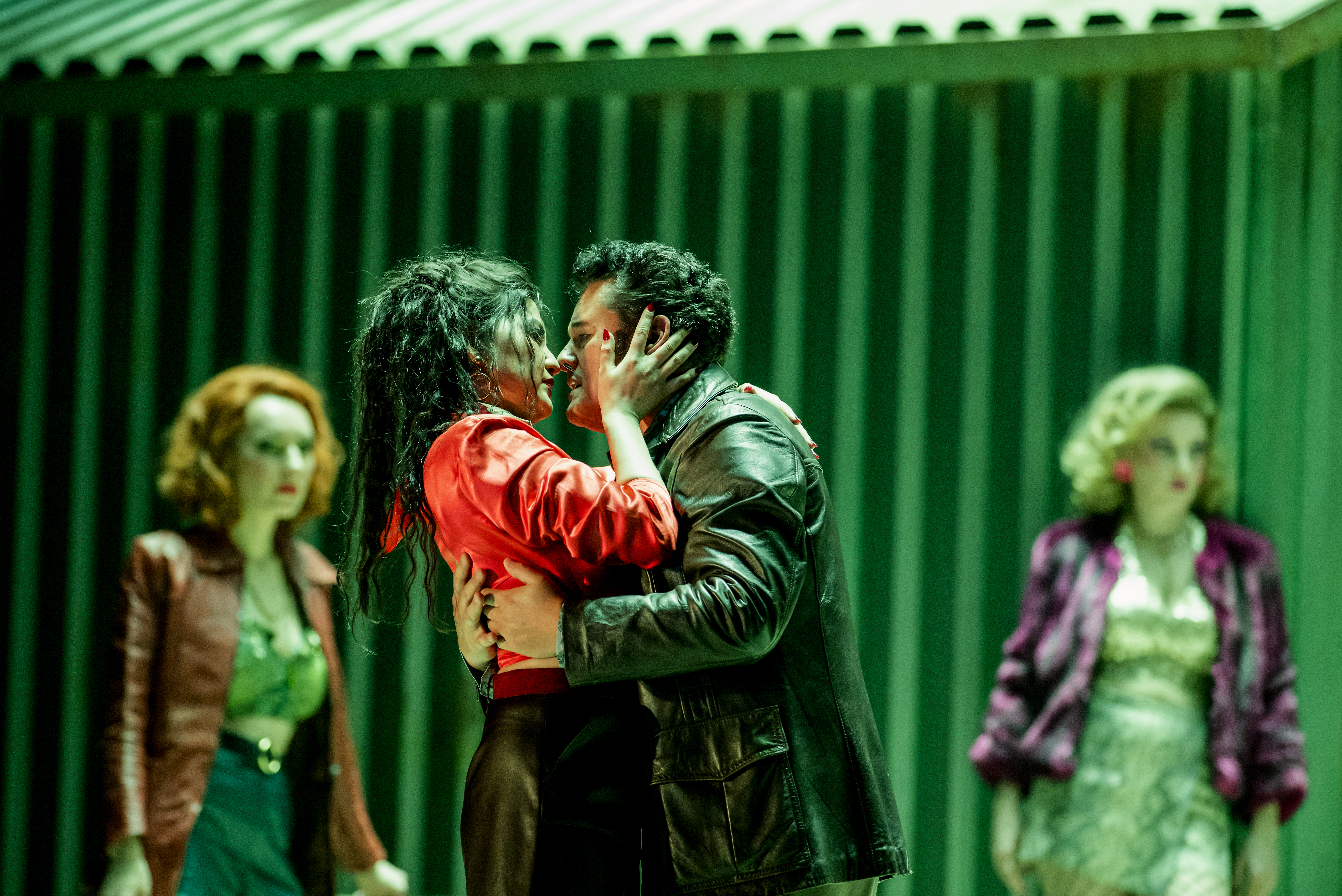 Two performers embrace passionately on a green-lit stage. Behind them are two other performers looking on. They are performing in The Royal Opera's production of Carmen. (6 of 12)