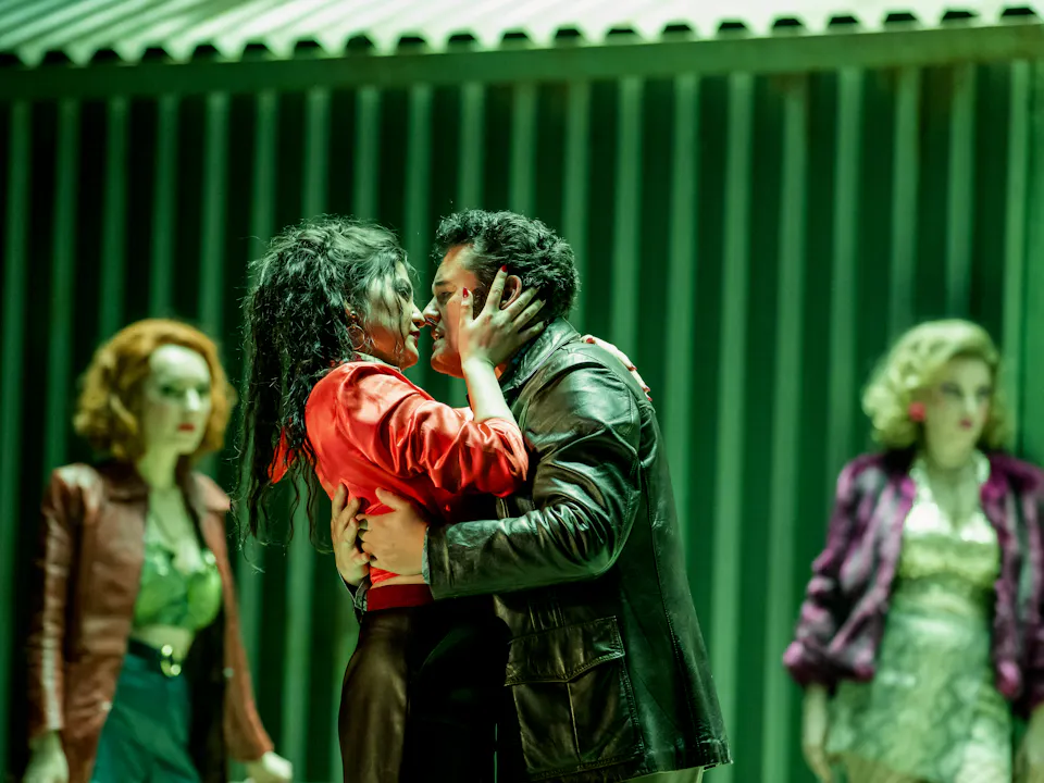 Two performers embrace passionately on a green-lit stage. Behind them are two other performers looking on. They are performing in The Royal Opera's production of Carmen.