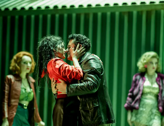 Two performers embrace passionately on a green-lit stage. Behind them are two other performers looking on. They are performing in The Royal Opera's production of Carmen. (6 of 12)