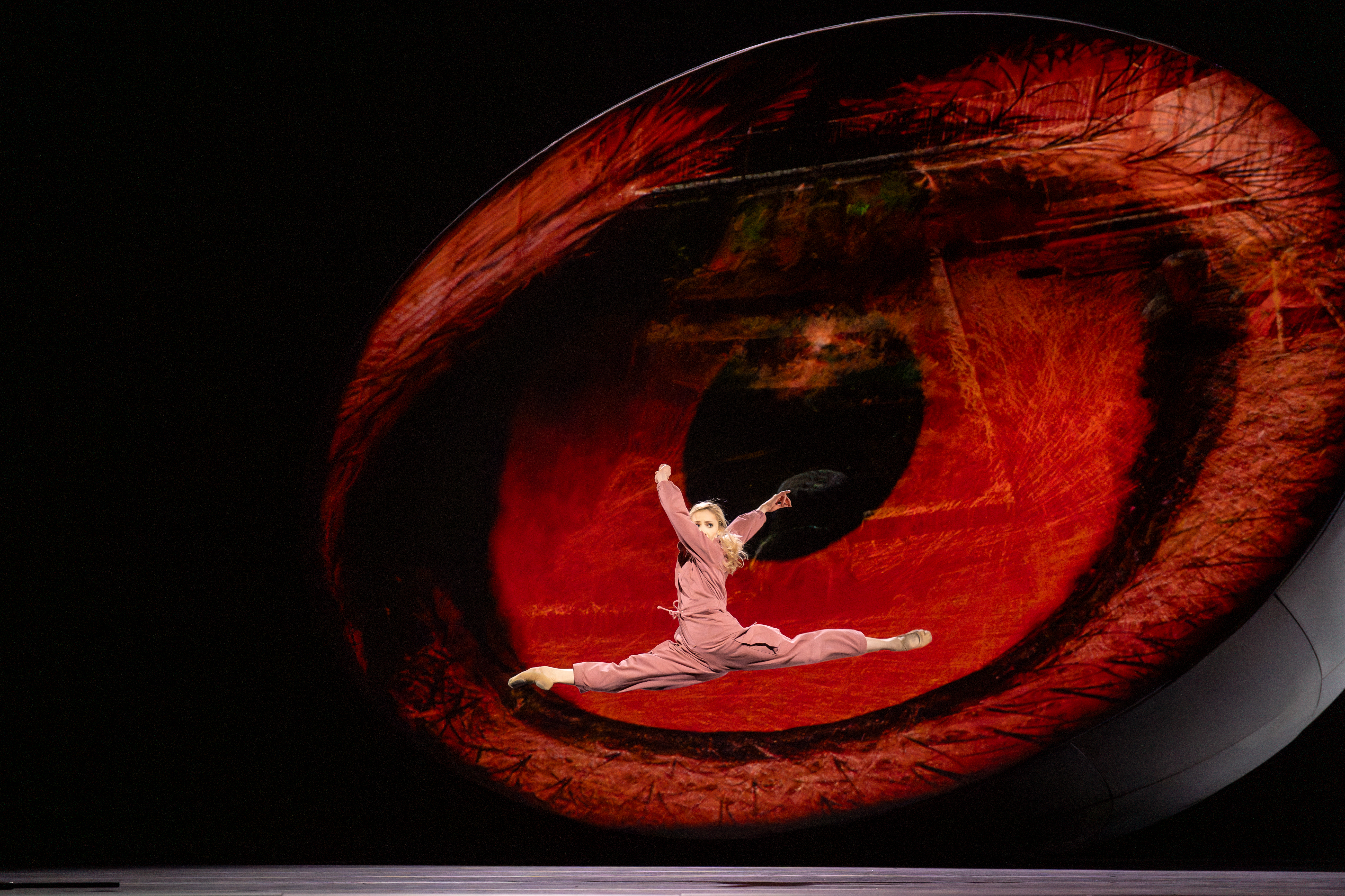 A ballet dancer wearing a pink jumpsuit launches themselves into the air in front of a backdrop of a big red eye. 