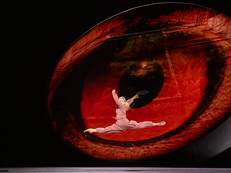 A ballet dancer wearing a pink jumpsuit launches themselves into the air in front of a backdrop of a big red eye.