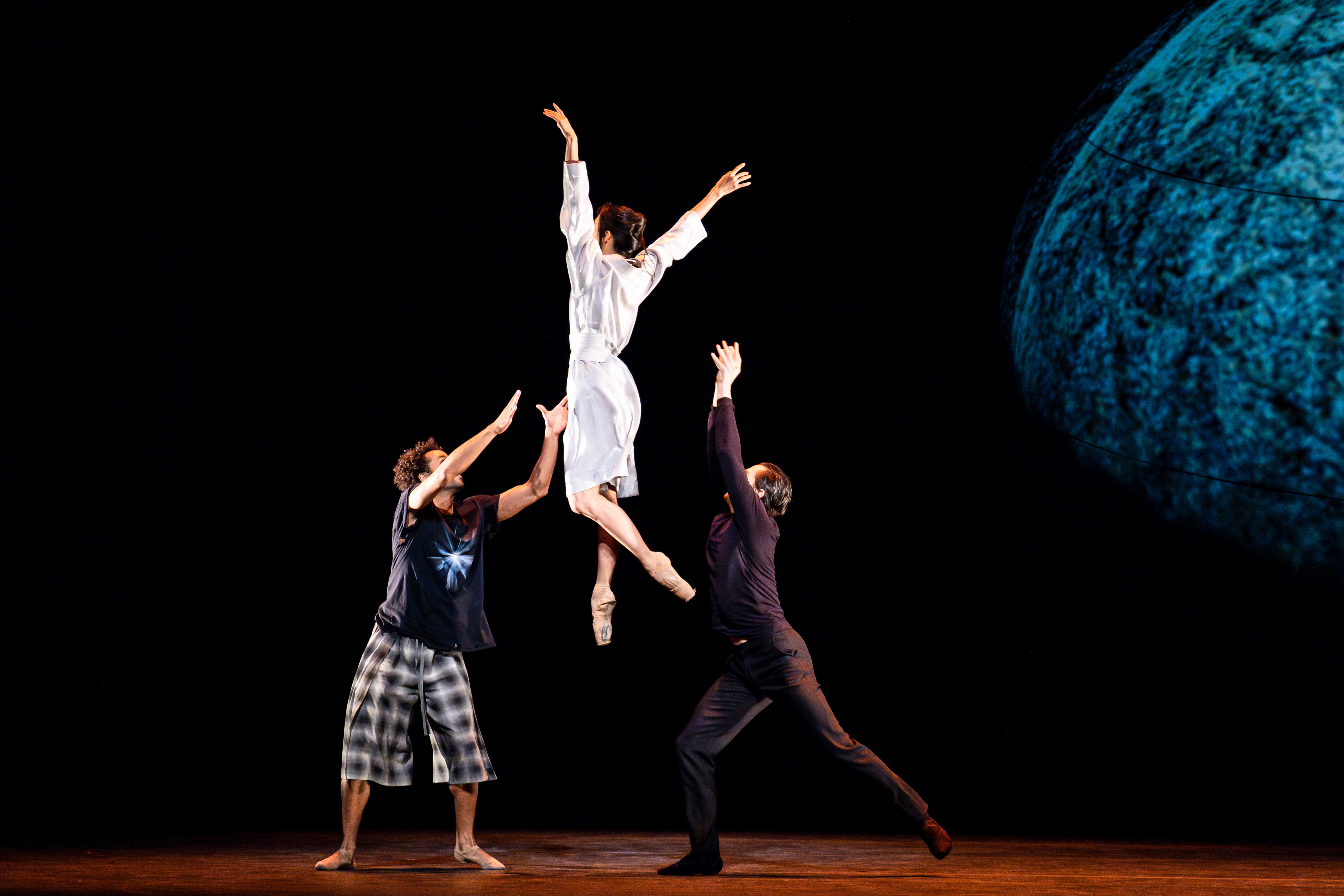 Three ballet dancers perform on stage. One dancer wearing all white launches themselves in the air while the other two hold their arms upwards as though to catch them. They perform in front of a dark backdrop. (1 of 7)