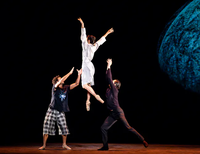Three ballet dancers perform on stage. One dancer wearing all white launches themselves in the air while the other two hold their arms upwards as though to catch them. They perform in front of a dark backdrop. (1 of 7)