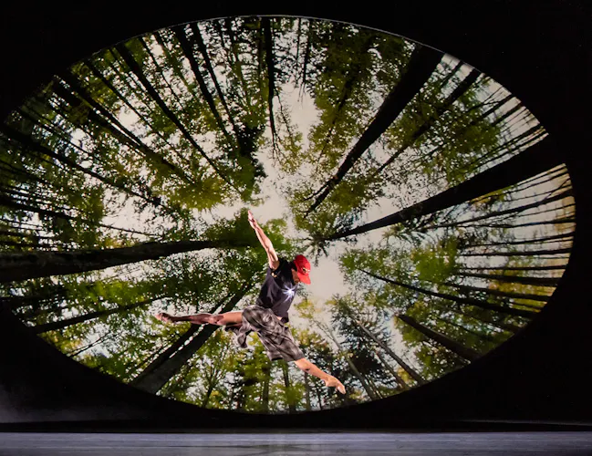 A ballet dancer wearing check patterned shorts launches themselves in to the air. They are performing in front of a dark backdrop with an image of a fish-eye lens view of a forest from below. (2 of 7)