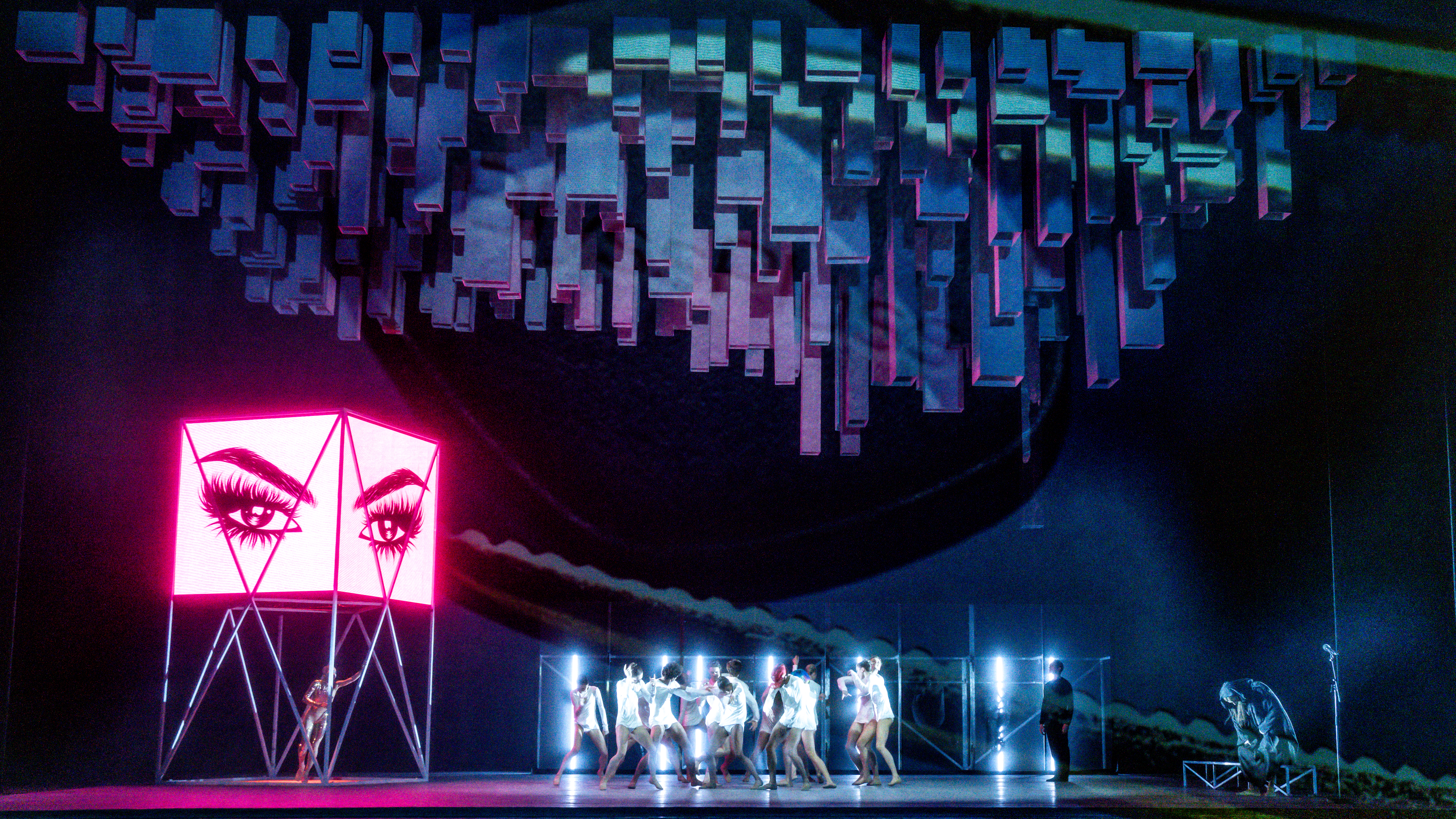 Several ballet dancers perform in the centre of a brightly neon lit stage all wearing white. Behind them are white neon strip lights. To the side of the stage is a pink lit advertising sign with an cartoon image of eyes and eyebrows. Geometric shapes  are lit from above. (4 of 7)