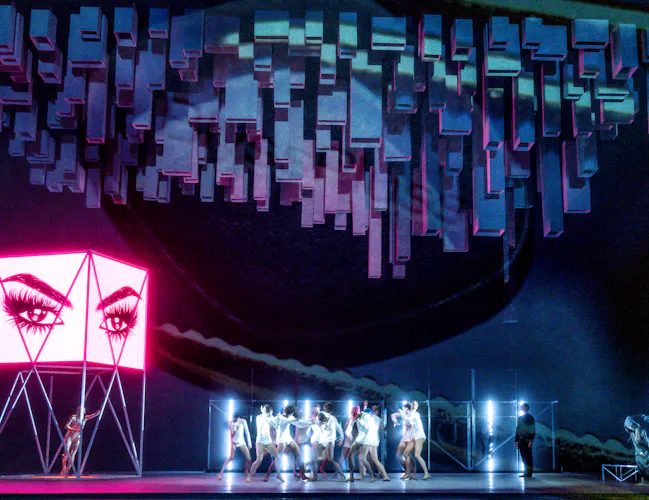 Several ballet dancers perform in the centre of a brightly neon lit stage all wearing white. Behind them are white neon strip lights. To the side of the stage is a pink lit advertising sign with an cartoon image of eyes and eyebrows. Geometric shapes are lit from above. (4 of 7)