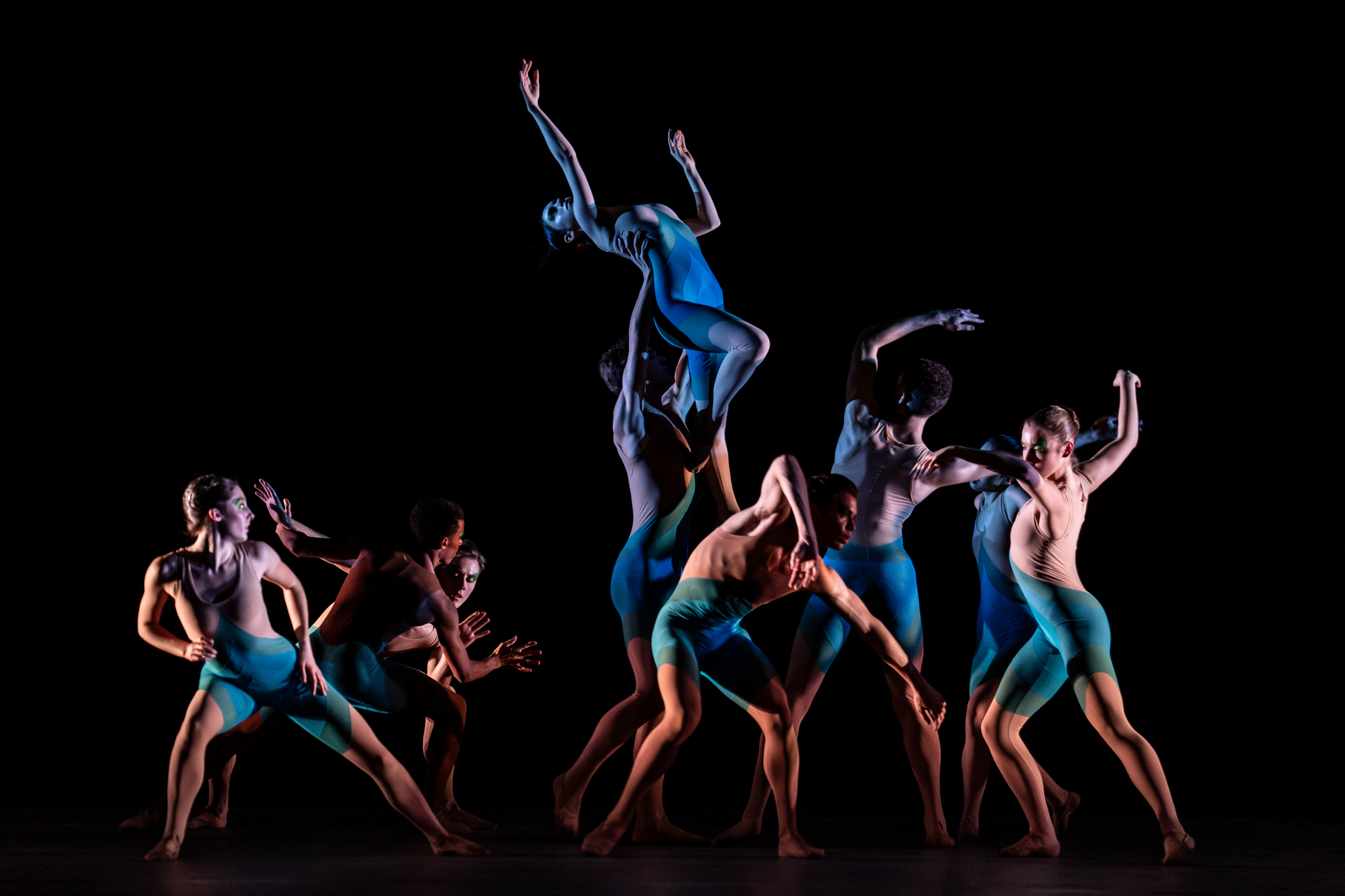 Several ballet dancers wearing blue leotards and short perform on a dark stage. One dancer is being held up by the group. (3 of 7)