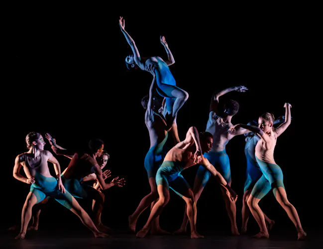 Several ballet dancers wearing blue leotards and short perform on a dark stage. One dancer is being held up by the group. (3 of 7)