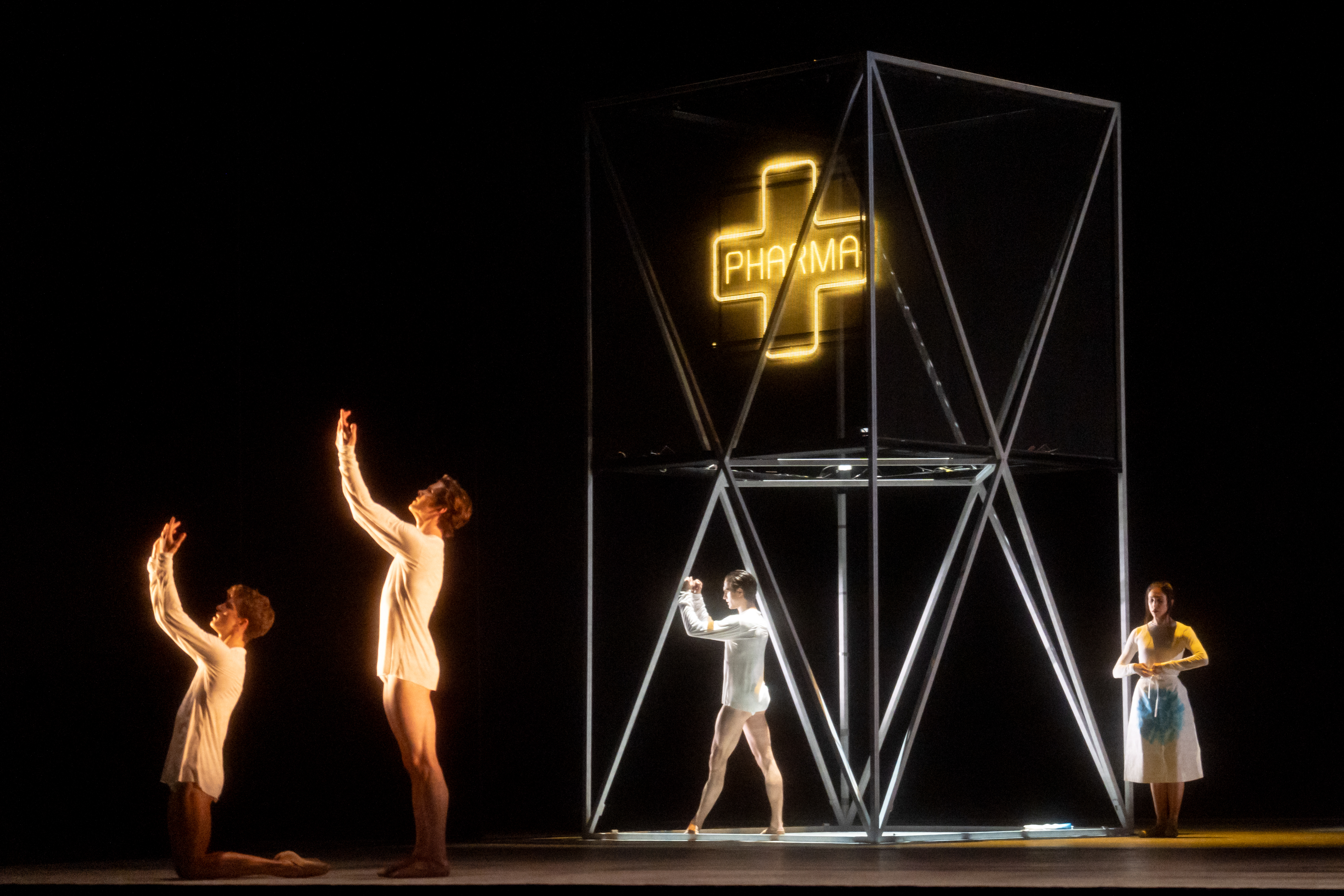 Four ballet dancers perform on a dark stage. In the centre of the stage is a metal scaffold with a neon lit pharmacy sign hanging down. (5 of 7)