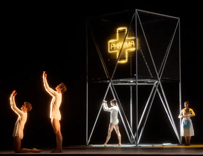 Four ballet dancers perform on a dark stage. In the centre of the stage is a metal scaffold with a neon lit pharmacy sign hanging down. (5 of 7)