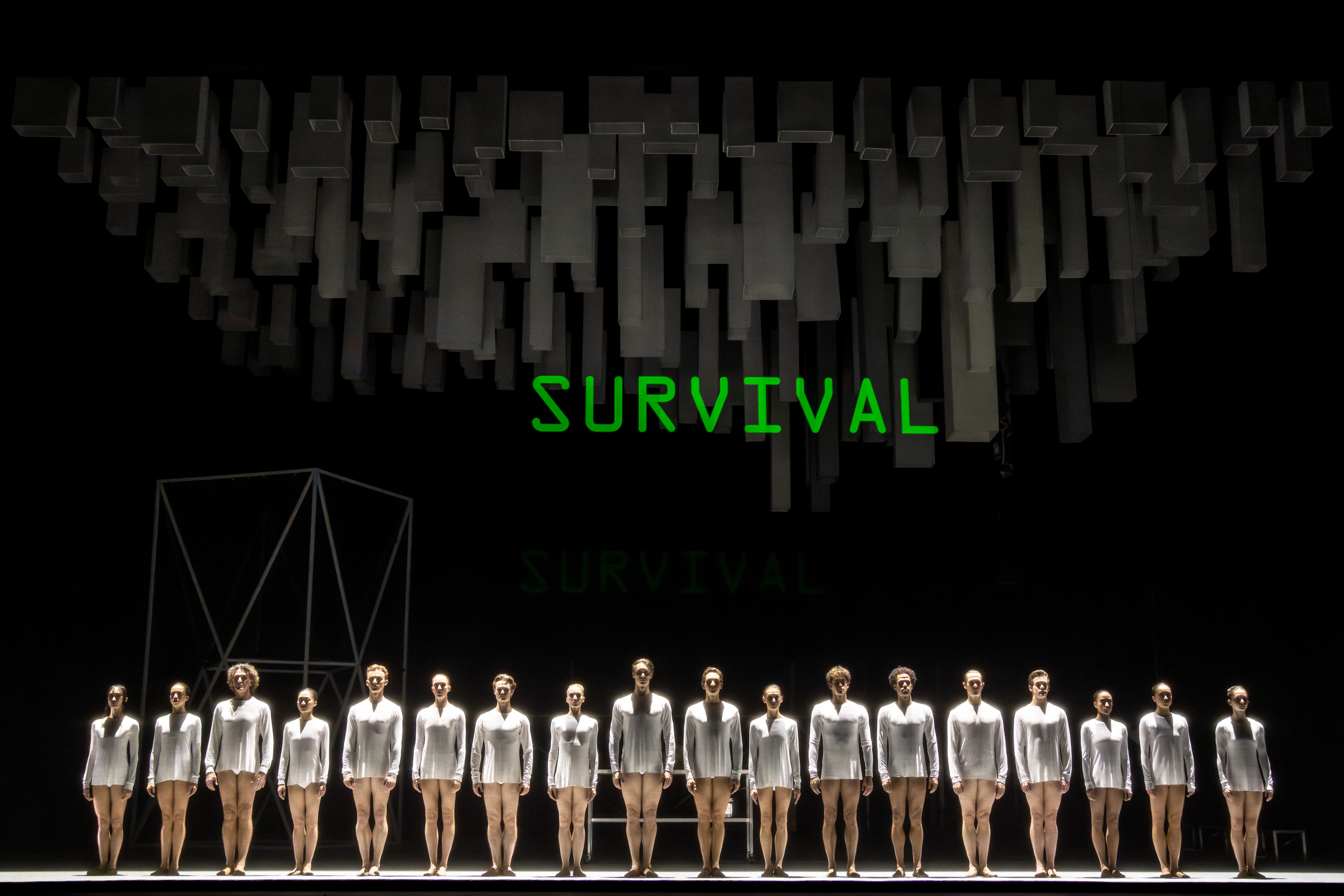 Several ballet dancers wearing short white costumes stand on a dark stage. Above them, them a neon lit sign reads "Survival". Above this sign, dark grey geometric shapes hang down, ominously over the performers. (6 of 7)
