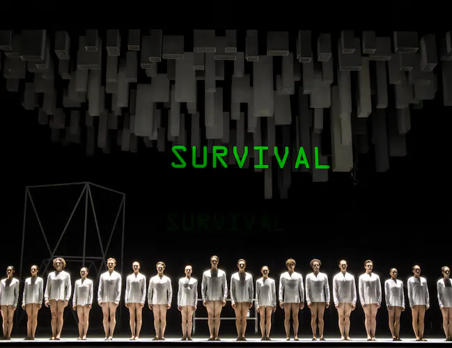 Several ballet dancers wearing short white costumes stand on a dark stage. Above them, them a neon lit sign reads "Survival". Above this sign, dark grey geometric shapes hang down, ominously over the performers. (6 of 7)