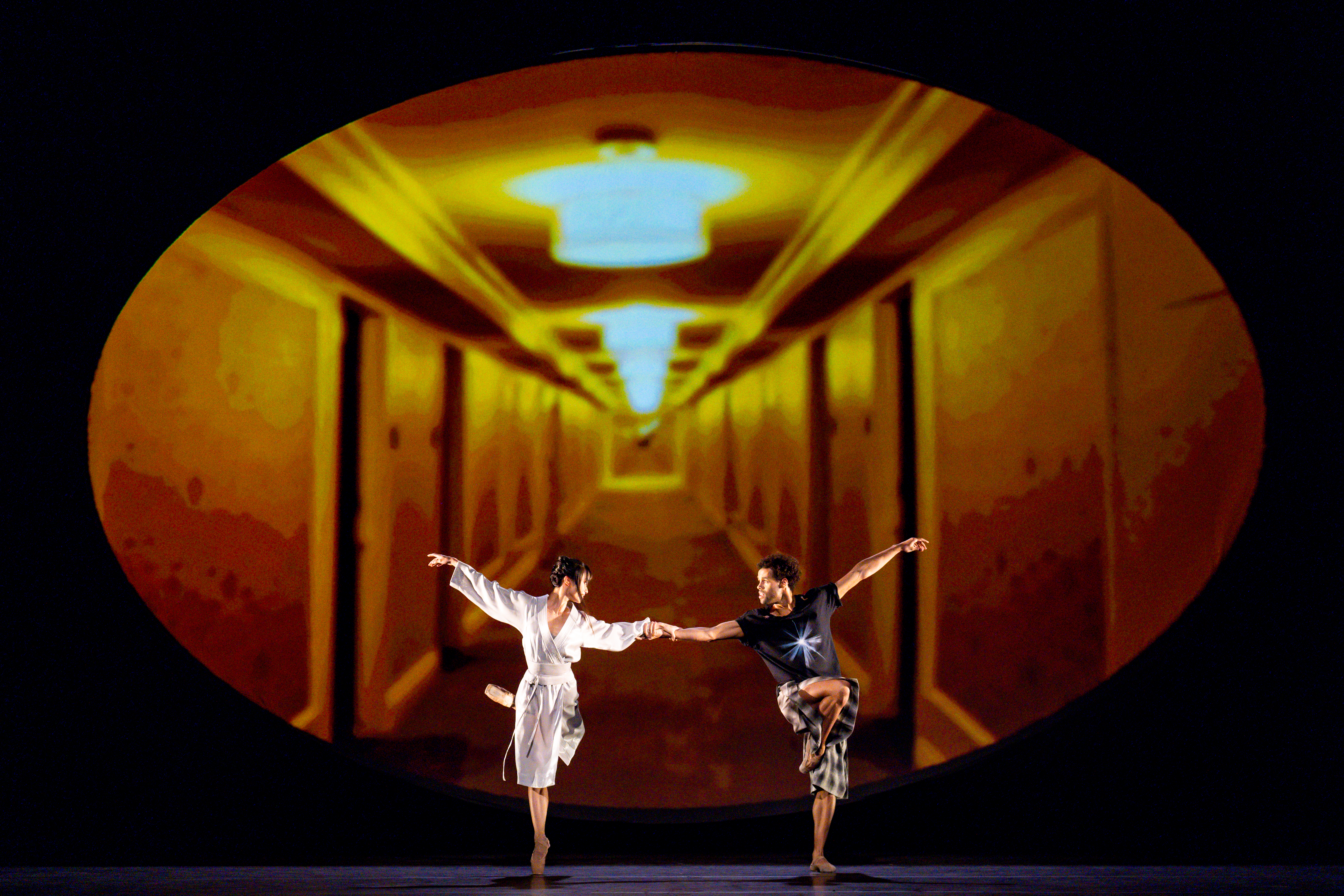 Two ballet dancers perform together on stage in front of dark backdrop with an oval framed image of an hallway that is lit bright orange. (7 of 7)