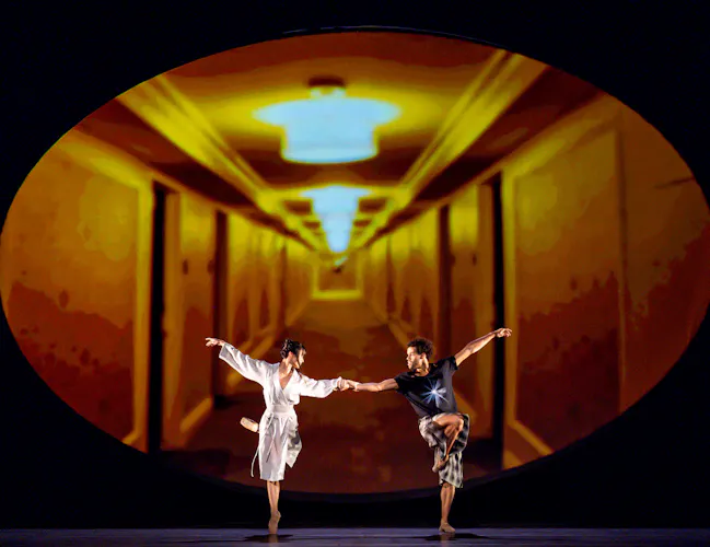 Two ballet dancers perform together on stage in front of dark backdrop with an oval framed image of an hallway that is lit bright orange. (7 of 7)