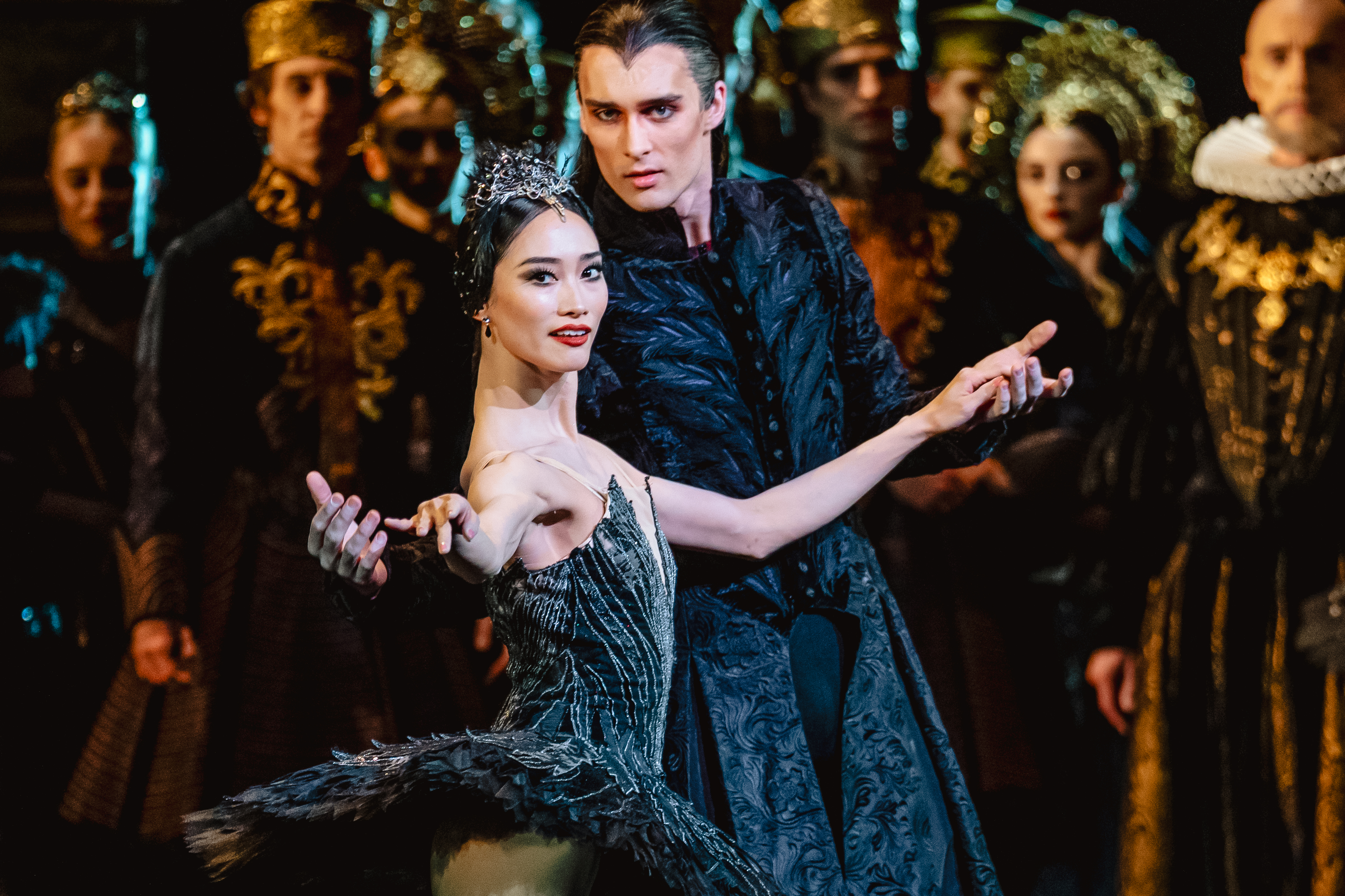 A ballet dancer wearing a black tutu and ornate tiara looks directly towards the audience. She is embraced from behind by another dancer wearing a long, embroidered dress jacket. Several dancers stand behind them on the stage. They are performing in The Royal Ballet's production of Swan Lake. (2 of 5)