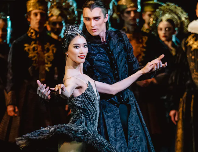 A ballet dancer wearing a black tutu and ornate tiara looks directly towards the audience. She is embraced from behind by another dancer wearing a long, embroidered dress jacket. Several dancers stand behind them on the stage. They are performing in The Royal Ballet's production of Swan Lake. (2 of 5)