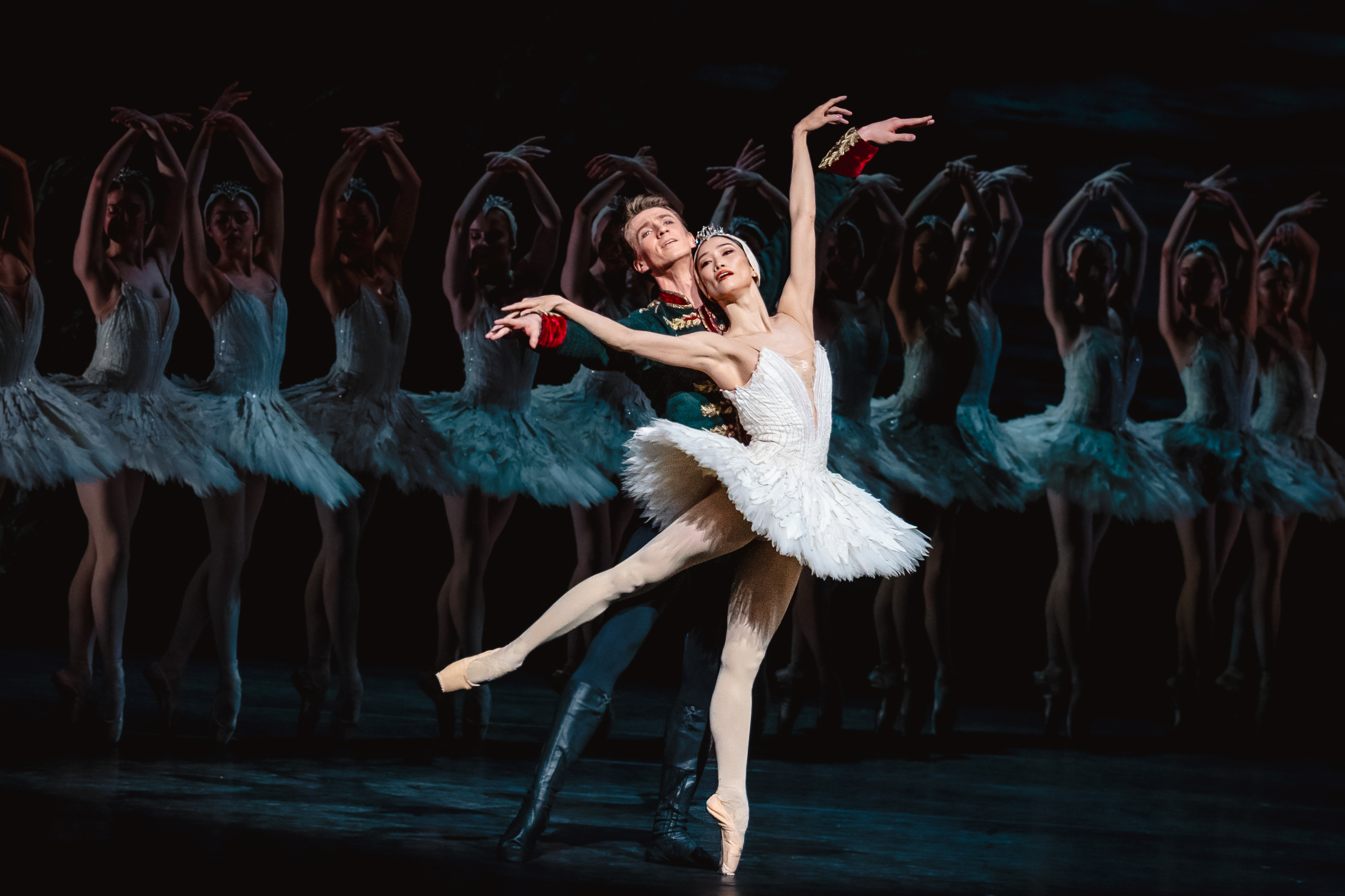 The Royal Ballet dancer Fumi Kaneko wears a white tutu and feathered headdress stands en pointe. She is embraced from behind by The Royal Ballet dancer Vadim Muntagirov who is wearing a black soldiers uniform. Slightly blurred from view, behind the two dancers, are a group of ballet dancers wearing the same white tutus and feathered headdresses. They are performing in The Royal Ballet's production of Swan Lake.
