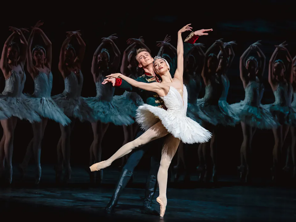 The Royal Ballet dancer Fumi Kaneko wears a white tutu and feathered headdress stands en pointe. She is embraced from behind by The Royal Ballet dancer Vadim Muntagirov who is wearing a black soldiers uniform. Slightly blurred from view, behind the two dancers, are a group of ballet dancers wearing the same white tutus and feathered headdresses. They are performing in The Royal Ballet's production of Swan Lake.