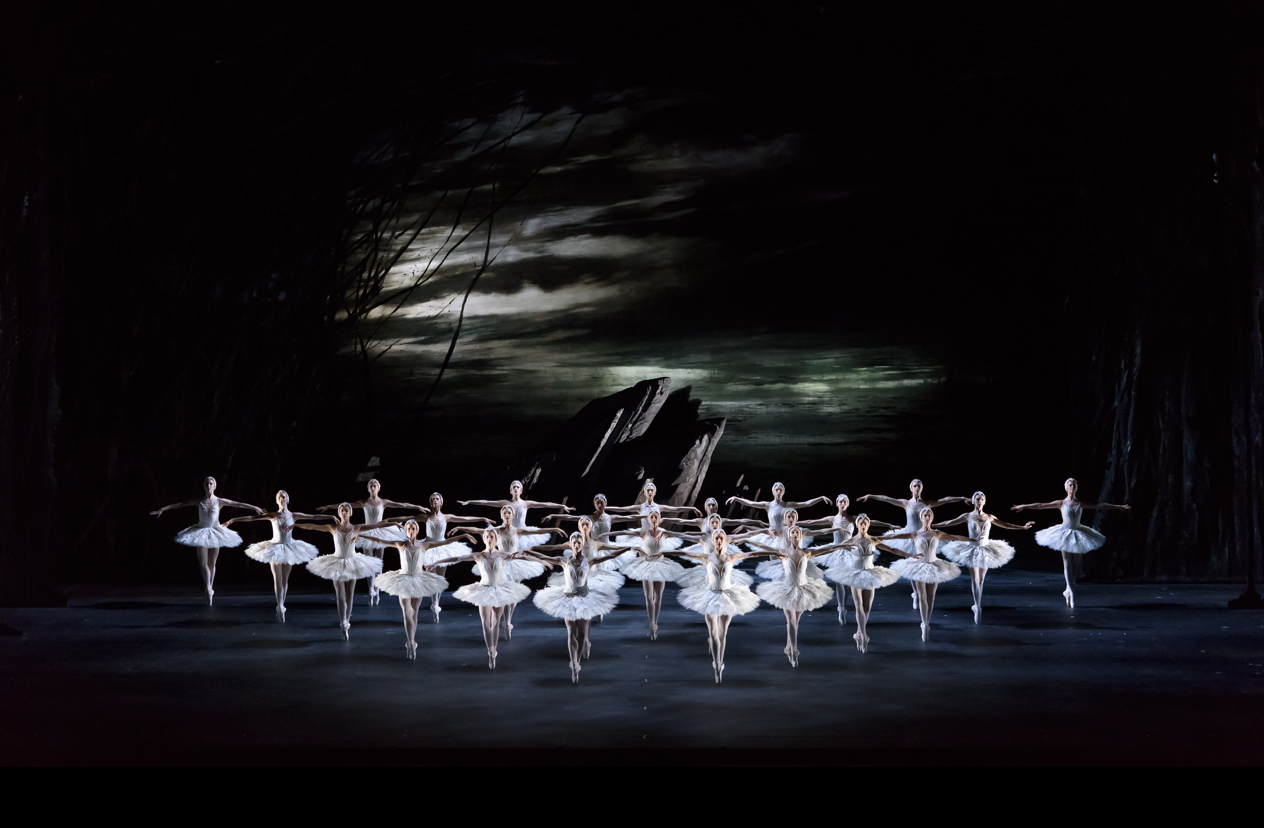 A view from above onto a dark set stage with several ballet dancers wearing white tutus and white feathered headdresses standing en pointe in a triangle shaped formation. They perform in front of a dark rock front and the background is dark, moody skies. They are performing in The Royal Ballet's production of Swan Lake. (1 of 5)